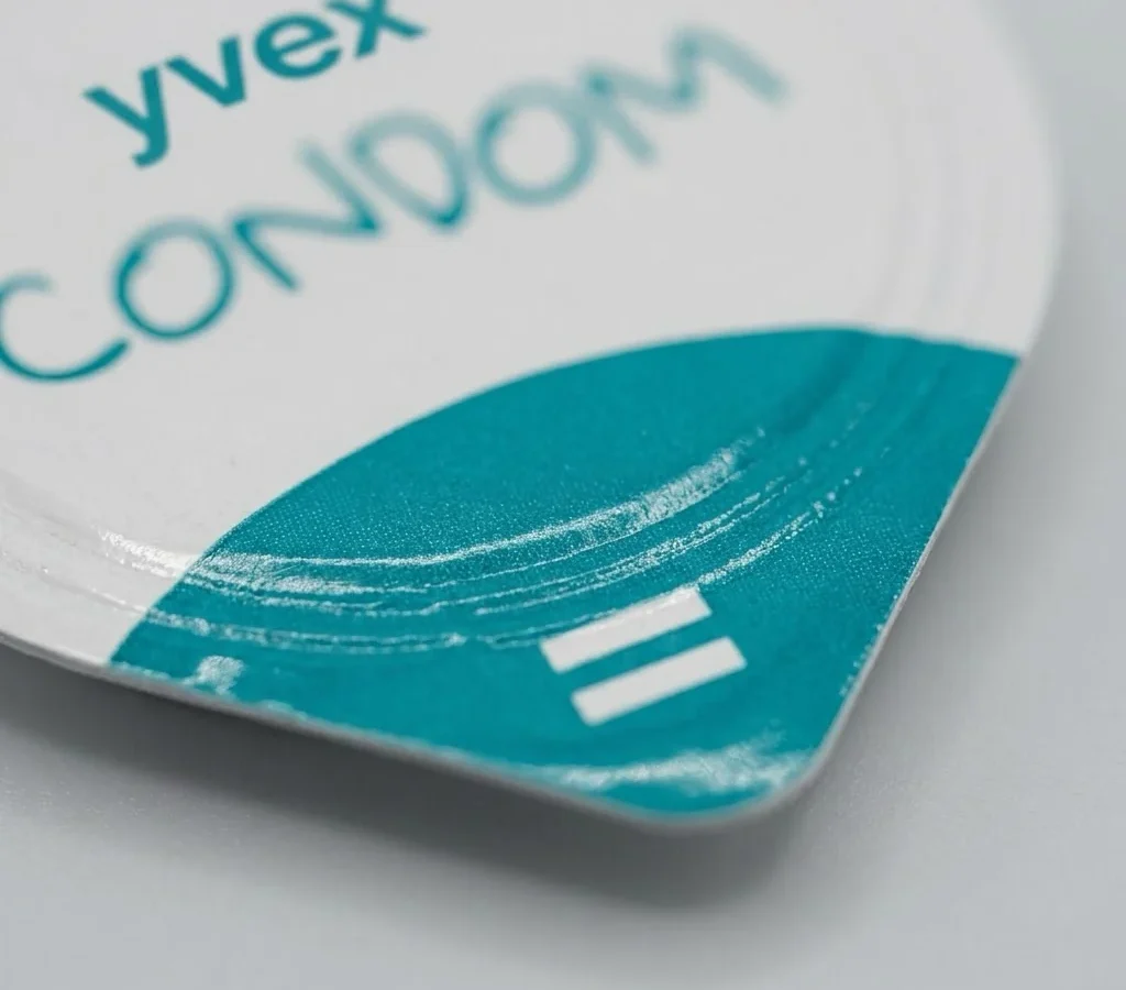 yvex condom core कैप का मैक्रो दृश्य: सफ़ेद फ़ॉइल पर टील रंग का ब्रश स्ट्रोक और अक्षर, अग्रभूमि में साफ़ उभरे हुए।