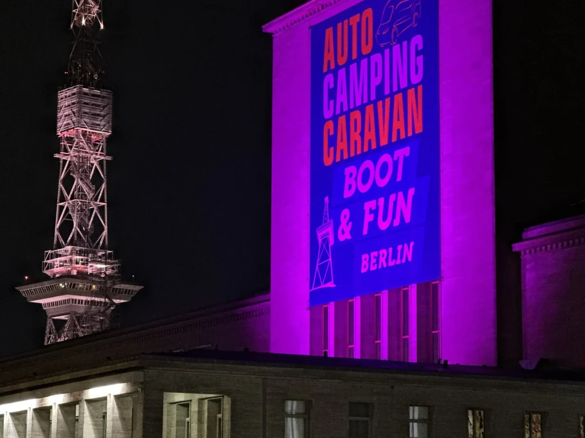 The illuminated entrance of a trade-fair hall at night, the event lettering in magenta light on the facade.