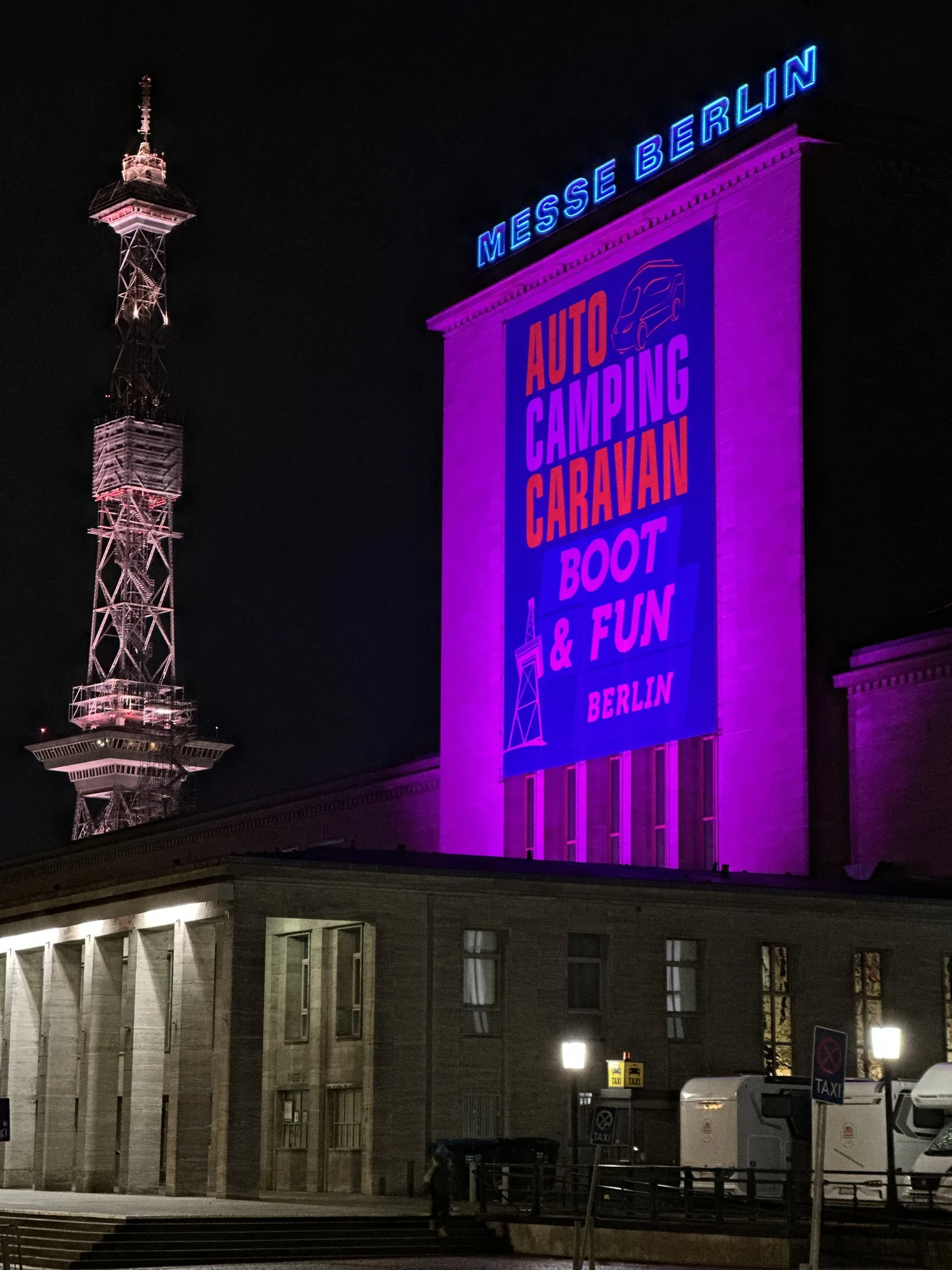 The illuminated entrance of a trade-fair hall at night, the event lettering in magenta light on the facade.