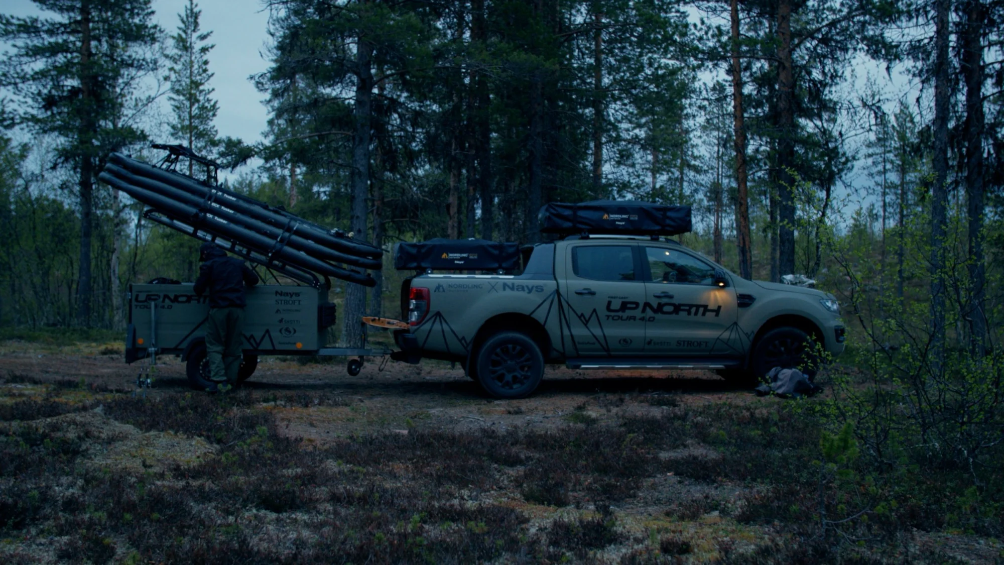 A pickup carrying a Nordling Outdoor rooftop tent and trailer parked on a track between pine trees.