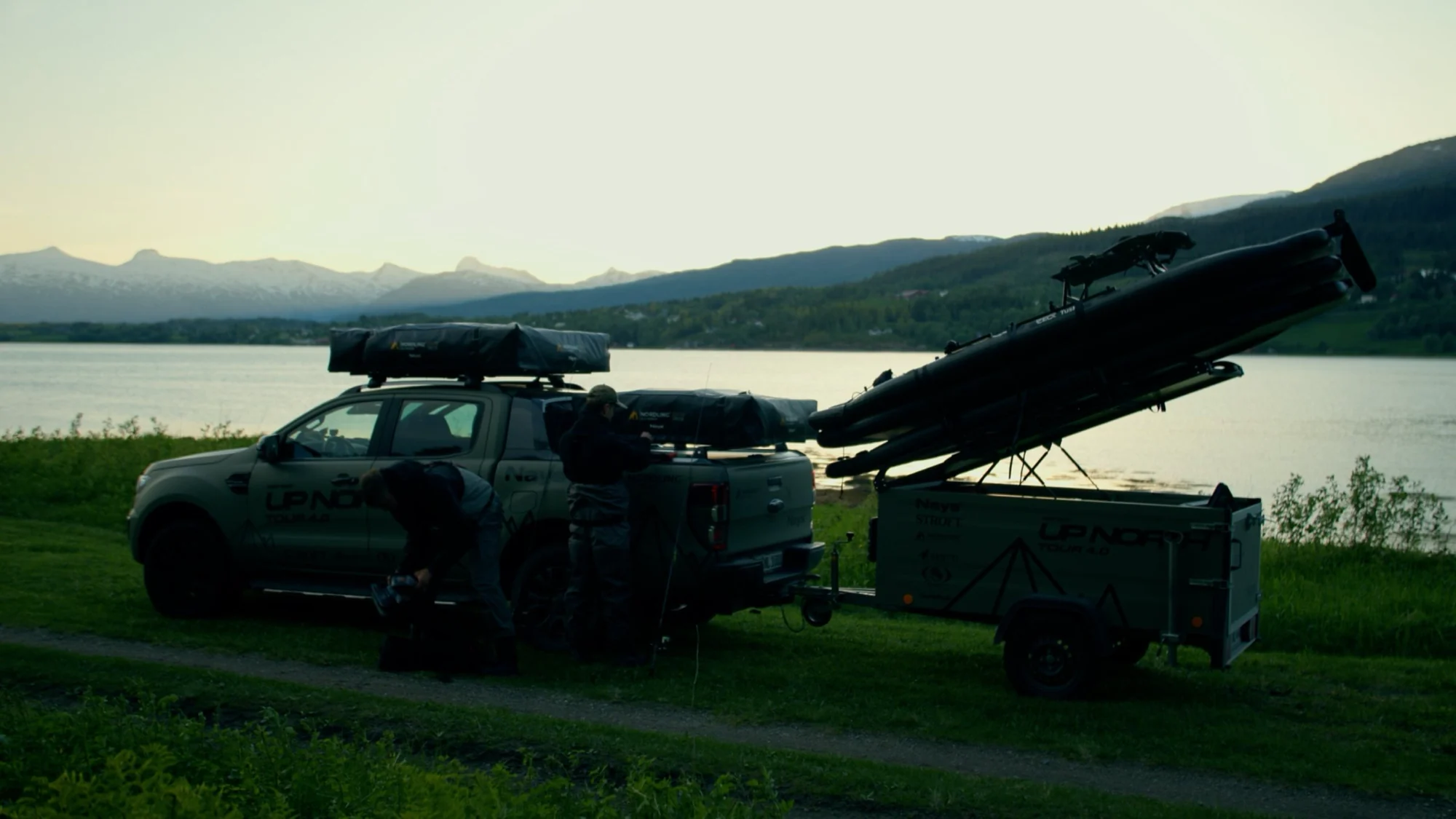 A pickup with a Nordling Outdoor rooftop tent and awning parked at a lakeside at dusk, hills across the water.