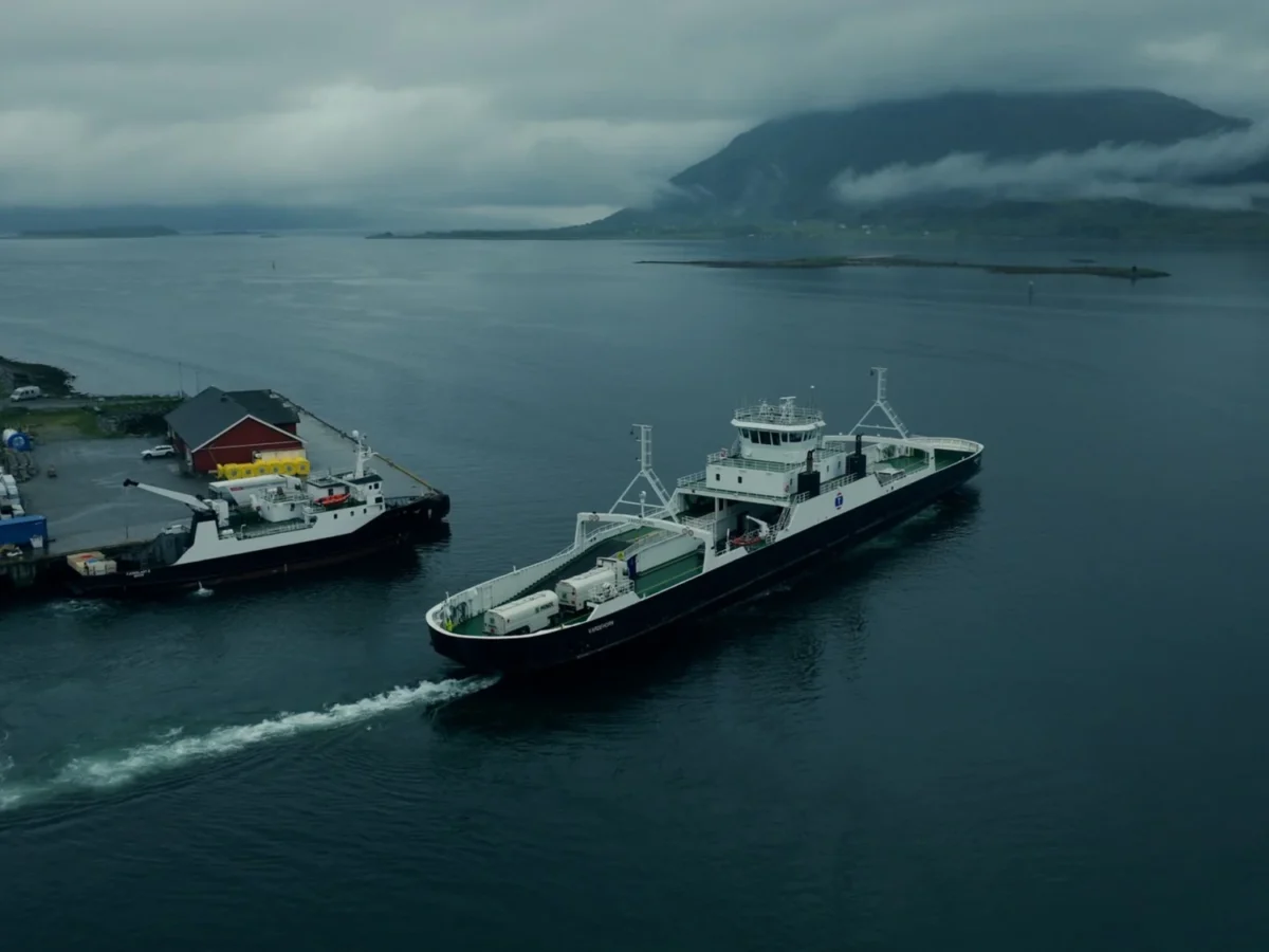 A ferry leaves a small harbour on a grey fjord with cloud-covered mountains behind, seen from above.