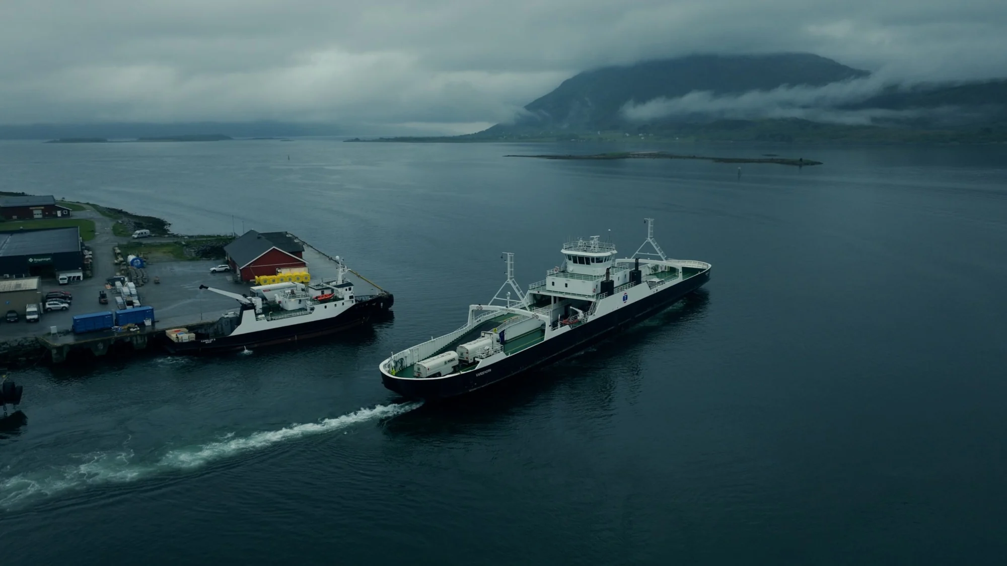 A ferry leaves a small harbour on a grey fjord with cloud-covered mountains behind, seen from above.