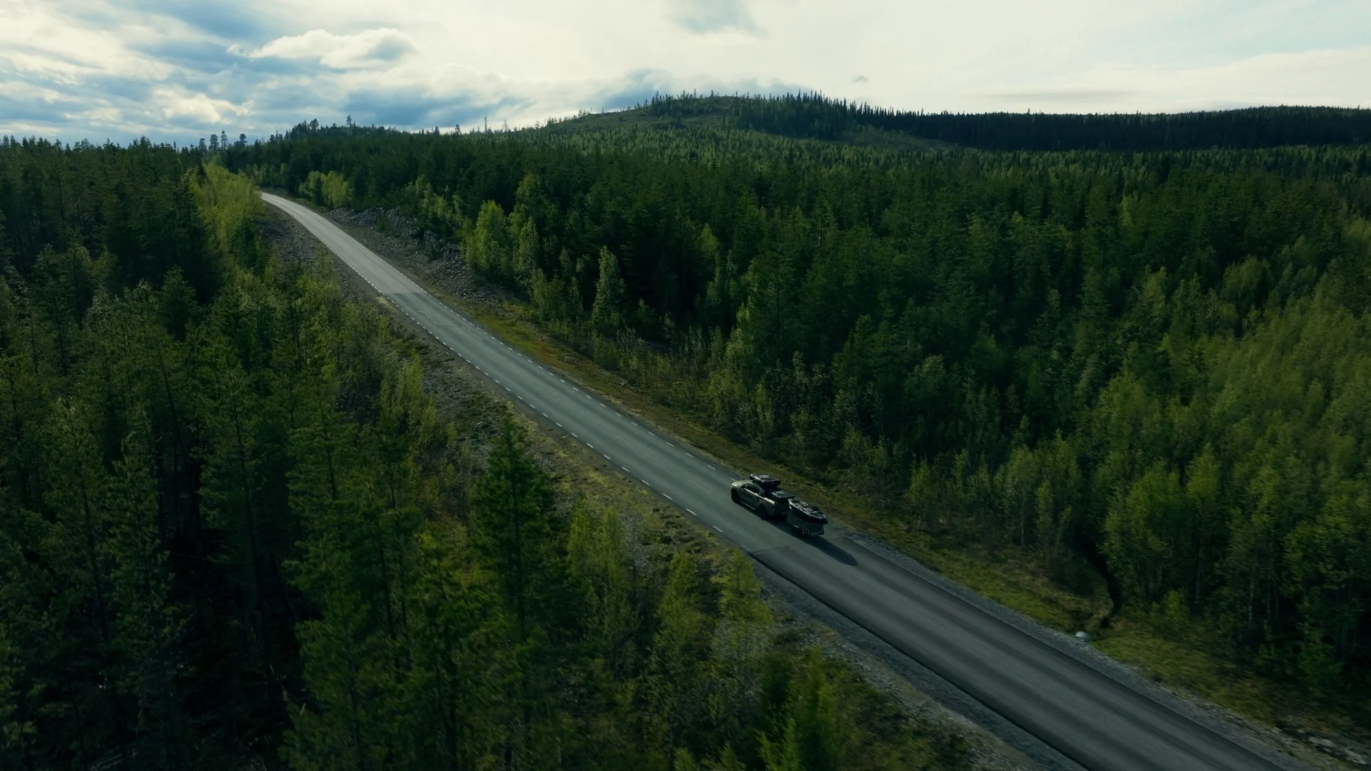 Drone view of a single road cutting through a Norwegian forest at dusk, the Green Beast small on the tarmac, hills and a fjord behind.