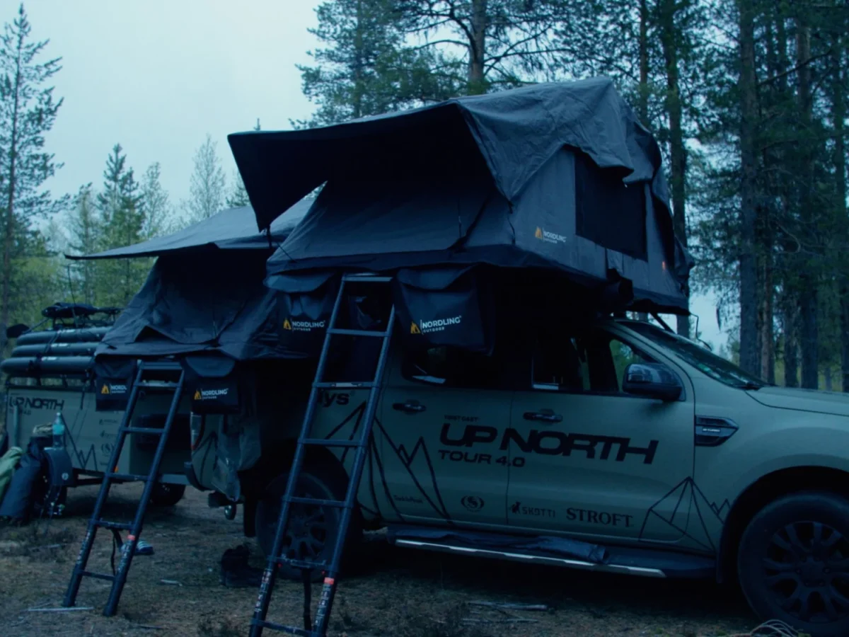 The Green Beast and its trailer with both Nordling rooftop tents open under tall pines, the UPNORTH print on the tent walls.