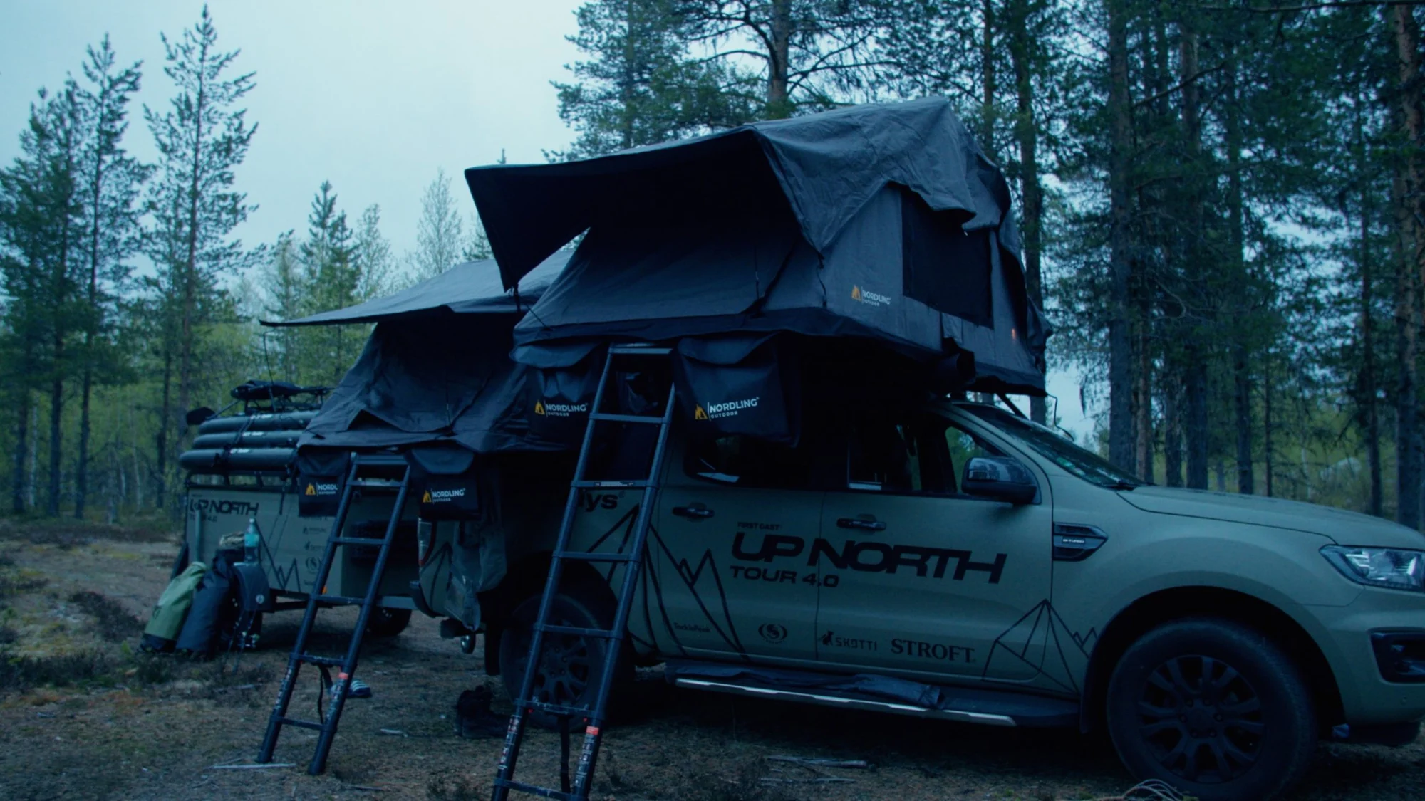 The Green Beast and its trailer with both Nordling rooftop tents open under tall pines, the UPNORTH print on the tent walls.