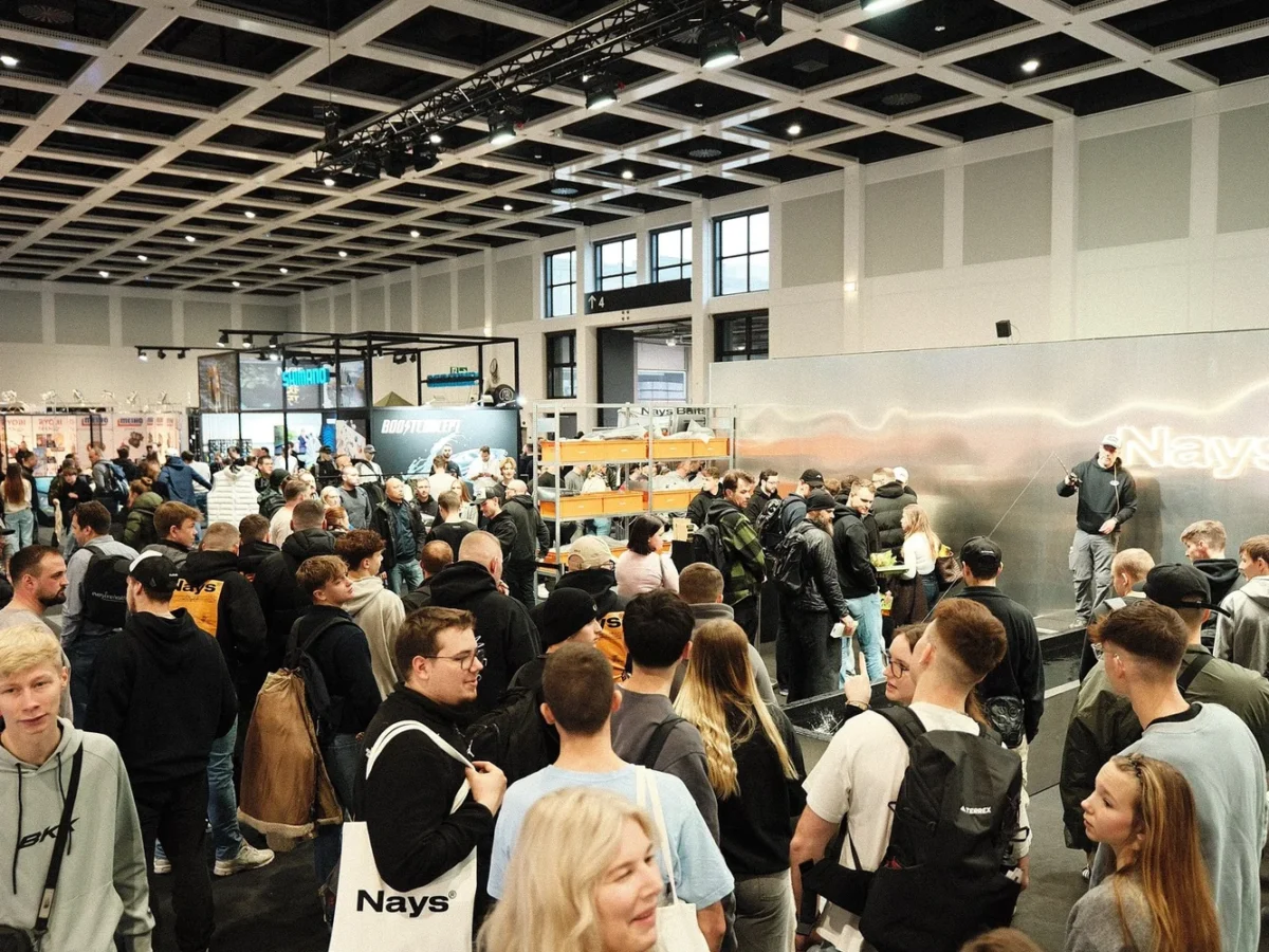 A crowd in front of the NAYS stand at a fishing fair, seen from above through the hall's steel lattice.
