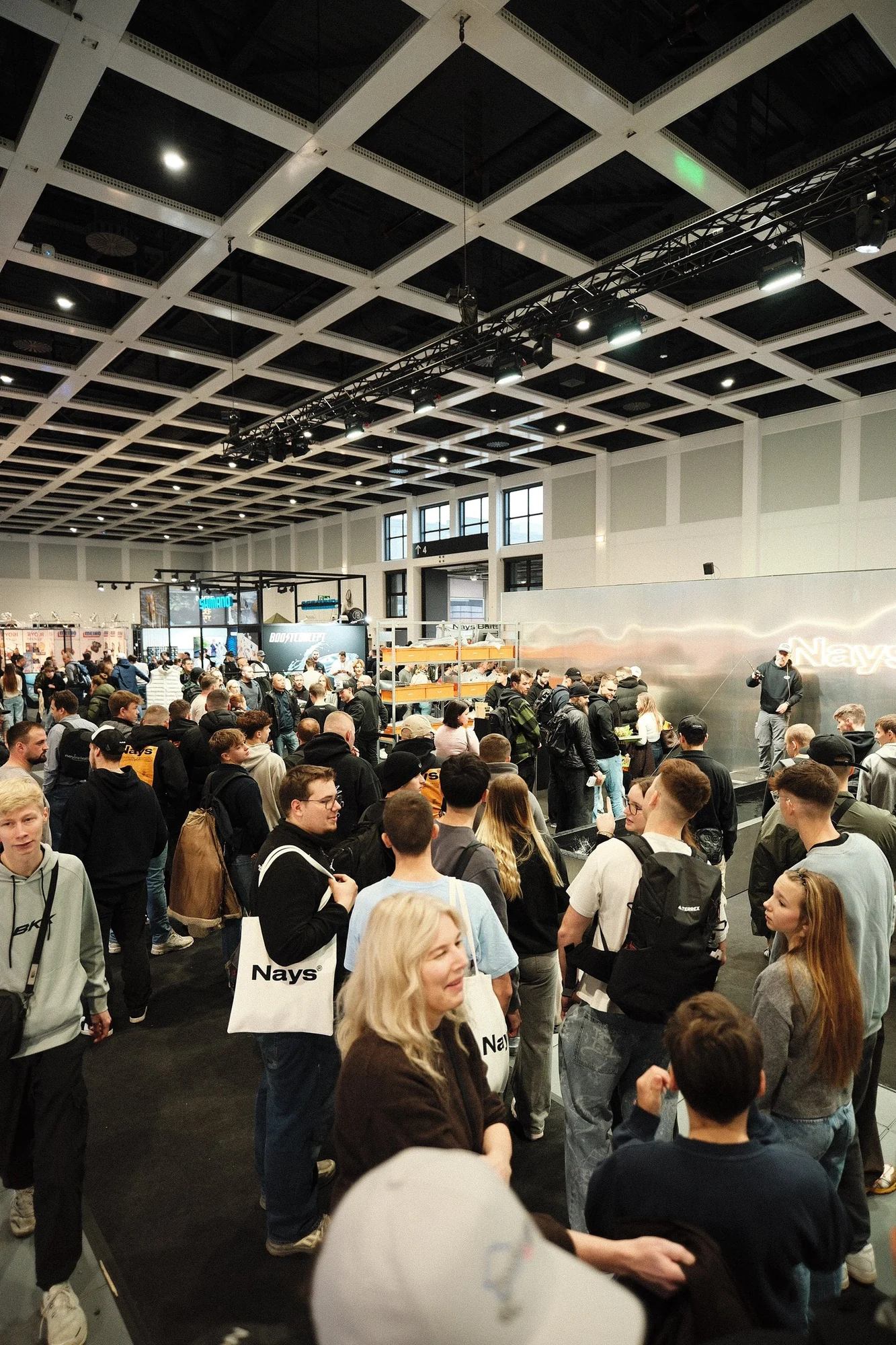 A crowd in front of the NAYS stand at a fishing fair, seen from above through the hall's steel lattice.