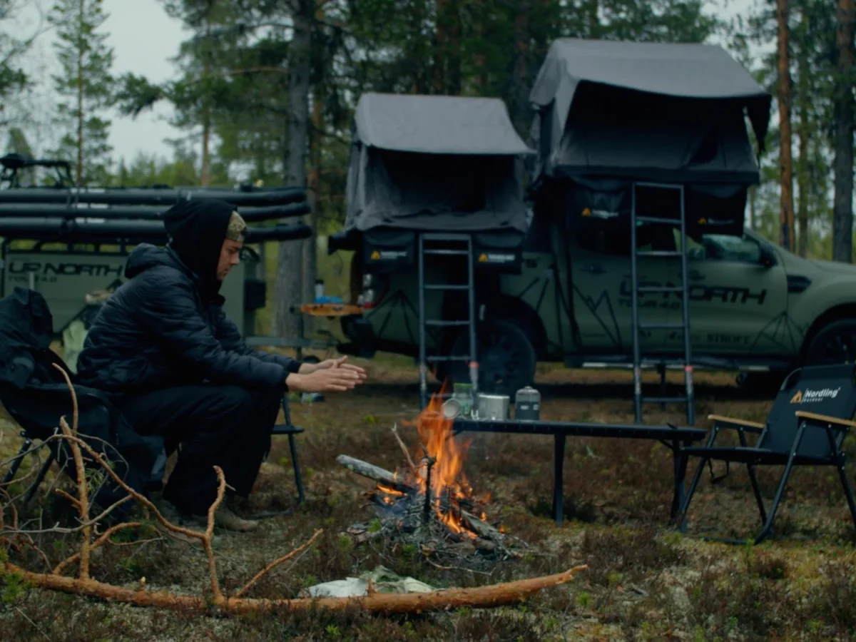 A man tends a camp fire at dusk beside the Green Beast pickup with its Nordling rooftop tent open, Nordling camping chairs around the fire.