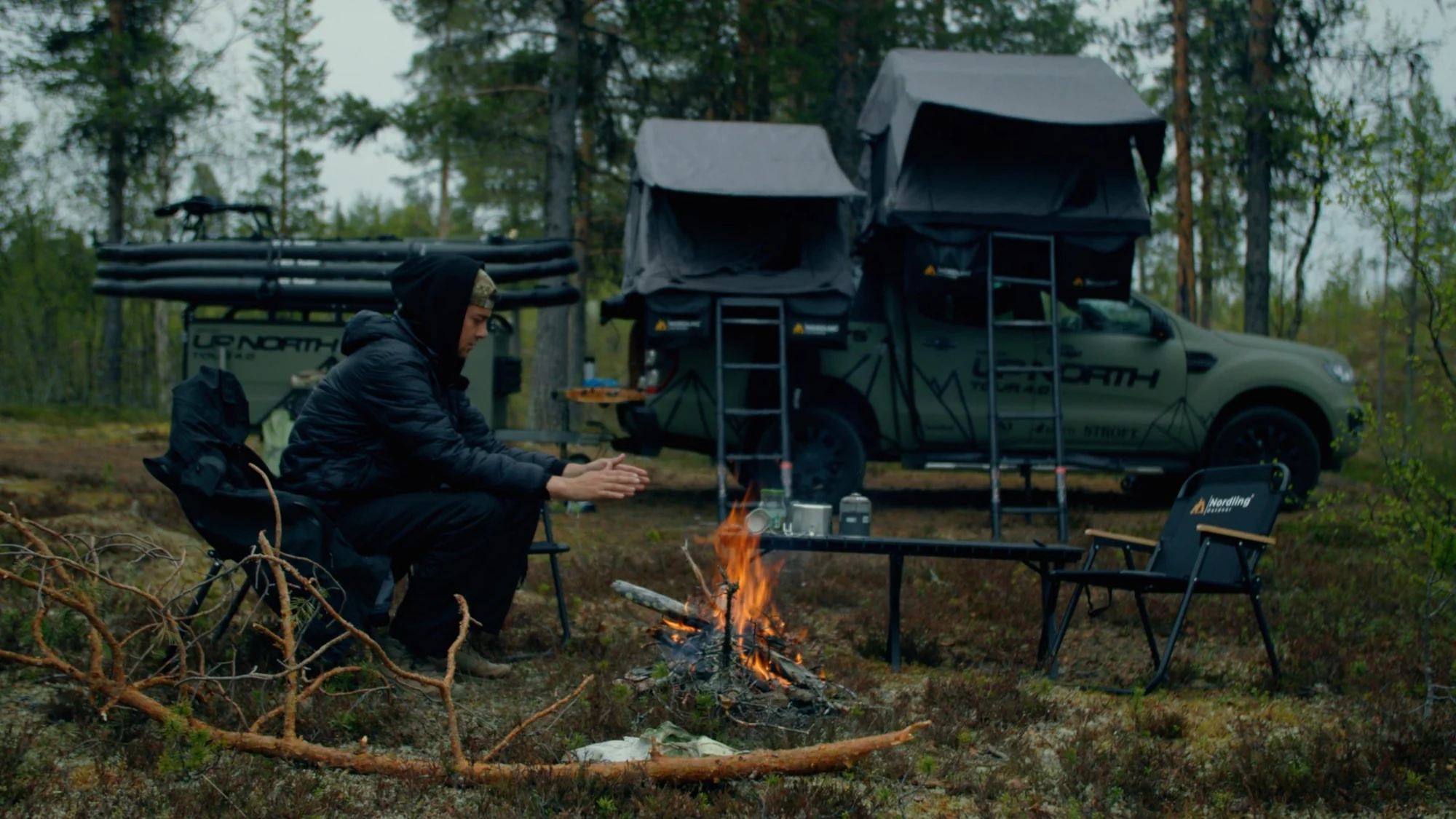 A man tends a camp fire at dusk beside the Green Beast pickup with its Nordling rooftop tent open, Nordling camping chairs around the fire.