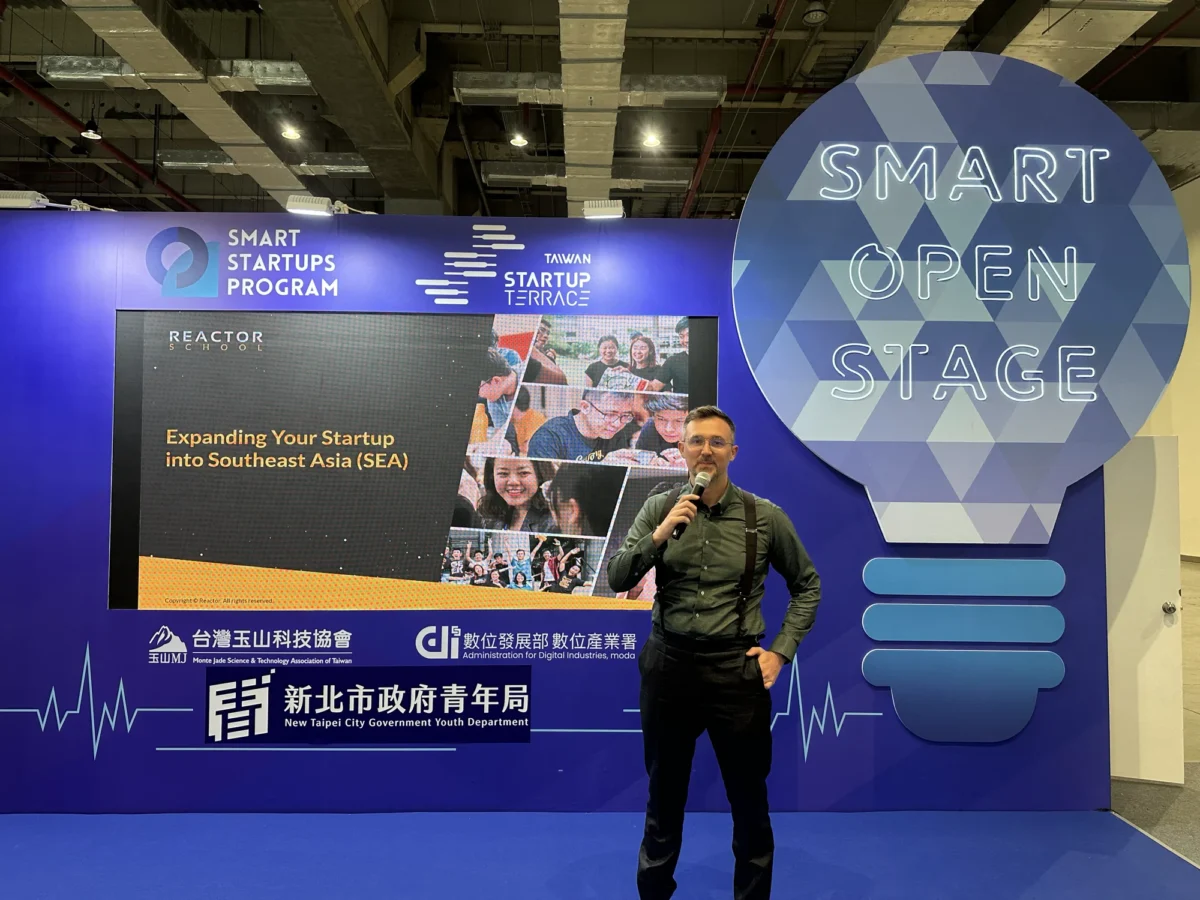 متحدث يحمل ميكروفوناً على منصة Smart Open Stage في فعالية لبرنامج Smart Startups Program، وخلفه خلفية على شكل مصباح ولافتات البرنامج.