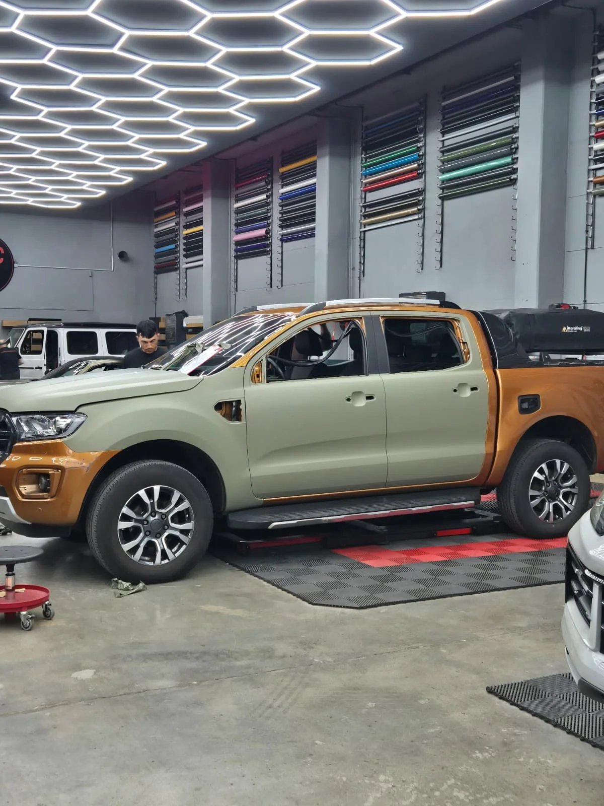 The pickup in a workshop under hexagon lights, half still orange and half wrapped in the olive green of UPNORTH.